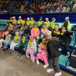Grupa dzieci siedzi na trybunach stadionu, w tle widoczna publiczność