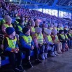 Grupa osób w kamizelkach odbezpieczających siedzi na trybunach stadionu