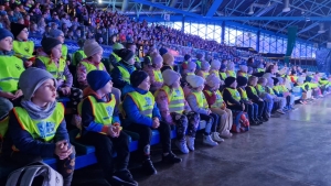 Grupa osób w kamizelkach odbezpieczających siedzi na trybunach stadionu