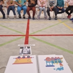 Grupa osób siedzi na krzesłach wokół kolorowych linii na podłodze