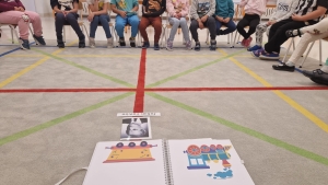 Grupa osób siedzi na krzesłach wokół kolorowych linii na podłodze