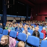 Grupa dzieci siedzi na niebieskich fotelach w dużej sali