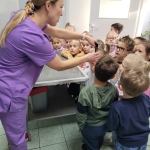 Kobieta w fioletowych ubraniach pokazuje dzieciom coś na stole