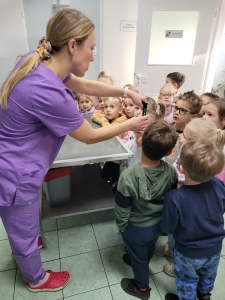 Kobieta w fioletowych ubraniach pokazuje dzieciom coś na stole