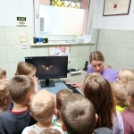 Grupa dzieci obserwuje komputer z nauczycielką w szkolnym pomieszczeniu