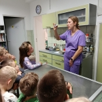 Kobieta w fioletowym stroju medycznym wyjaśnia coś dzieciom w gabinecie