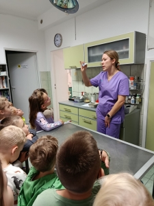 Kobieta w fioletowym stroju medycznym wyjaśnia coś dzieciom w gabinecie