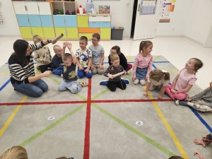 Grupa dzieci siedzi na podłodze wokół kobiety, która coś pokazuje