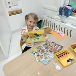 Dziewczynka siedzi przy stole i trzyma kolorowy rysunek z owocami