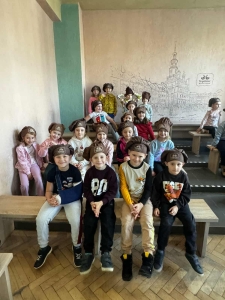 Grupa dzieci w czapkach siedzi na ławkach w pomieszczeniu