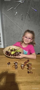 Dziewczynka trzyma koszyk z orzechami i kasztanami, na stole figurki z orzechów