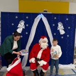 Santa Claus siedzi na krześle, dziecko trzyma prezent, kobieta rozdaje dary