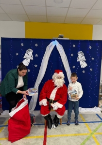 Santa Claus siedzi na krześle, dziecko trzyma prezent, kobieta rozdaje dary