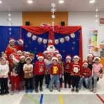 Grupa dzieci i dorosłych w czapkach mikołajkowych stoi przed dekoracją