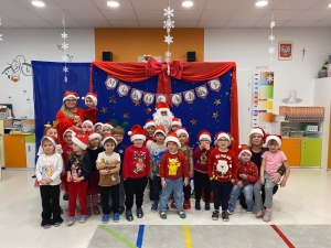 Grupa dzieci i dorosłych w czapkach mikołajkowych stoi przed dekoracją