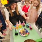 Kobieta w czarnym kapeluszu pokazuje dzieciom kolorowe butelki. (45 znaków)