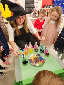 Kobieta w czarnym kapeluszu pokazuje dzieciom kolorowe butelki. (45 znaków)