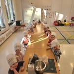 Dzieci w czapkach kuchennych siedzą przy długim stole w przedszkolu