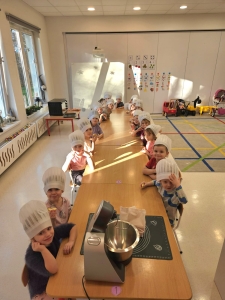 Dzieci w czapkach kuchennych siedzą przy długim stole w przedszkolu