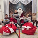 Dzieci siedzą z Mikołajem, trzymając prezenty. Na podłodze leżą worki