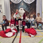 Dzieci siedzą z Mikołajem, który wręcza prezenty. Na podłodze leży worki